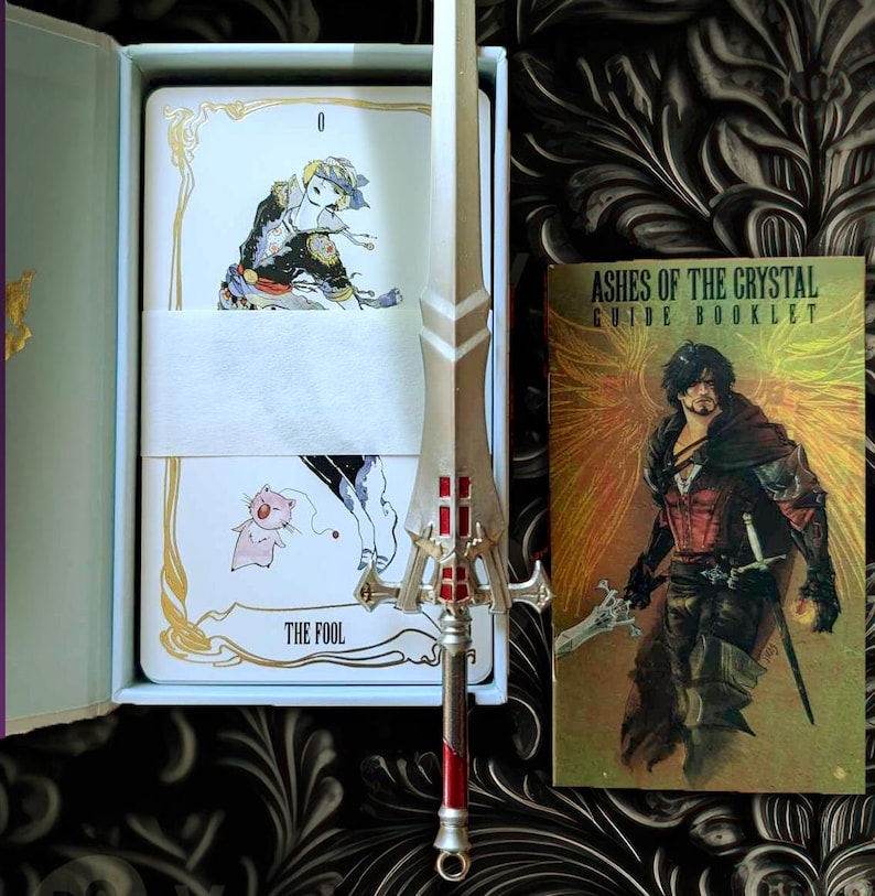 May include: A set of tarot cards with a guide booklet and a silver sword. The tarot card shows a character with a pink creature. The card is labeled "The Fool". The guide booklet is titled "Ashes of the Crystal Guide Booklet".