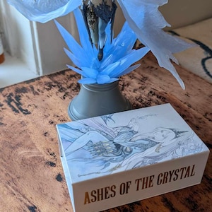 May include: A blue and white figurine of a woman with wings, standing on a crystal base. The figurine is on a wooden table next to a white box with the words "Ashes of the Crystal" printed in gold.