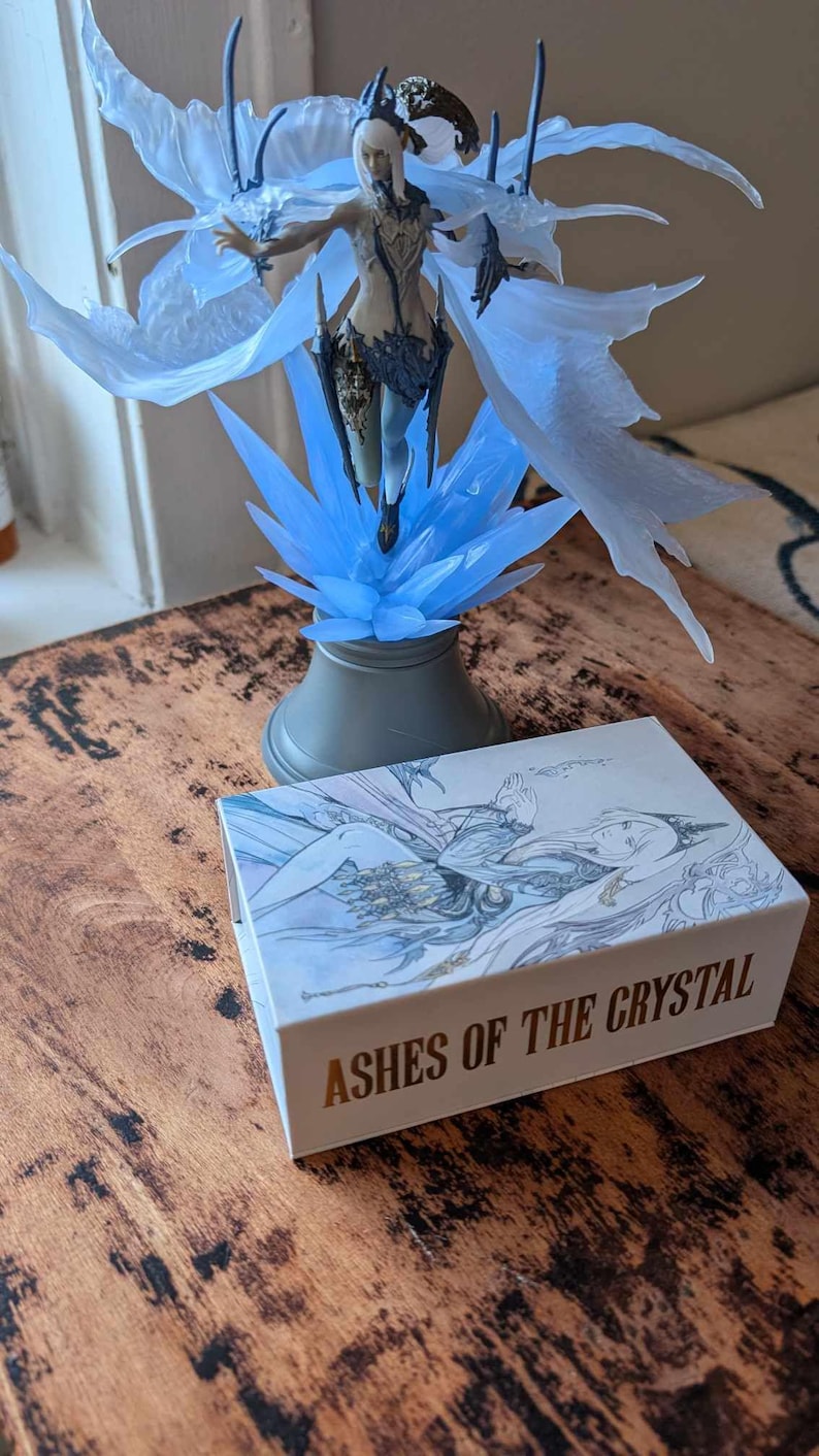 May include: A blue and white figurine of a woman with wings, standing on a crystal base. The figurine is on a wooden table next to a white box with the words "Ashes of the Crystal" printed in gold.