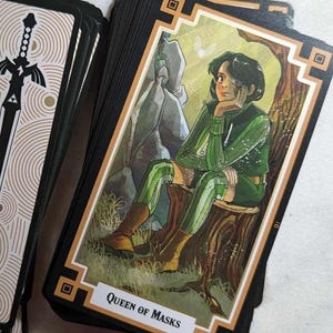 May include: A tarot card with a black and gold border. The card features a young person with dark hair sitting on a tree stump in a forest setting. The card is titled "Queen of Masks".