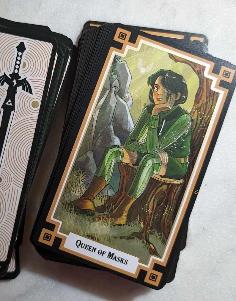 May include: A tarot card with a black and gold border. The card features a young person with dark hair sitting on a tree stump in a forest setting. The card is titled "Queen of Masks".
