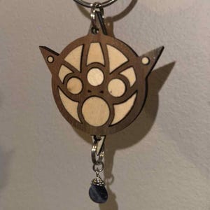 May include: A wooden keychain featuring a stylized cat face design with a light wood inlay. The keychain has a silver ring and a small, dark blue gemstone pendant. The cat face has pointed ears and circular features.