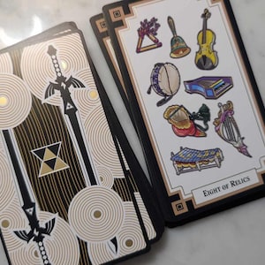 May include: Two tarot cards, one with a black and gold design featuring a sword and the other with a white background and colorful illustrations of musical instruments. The card with the musical instruments has the text "Eight of Relics".