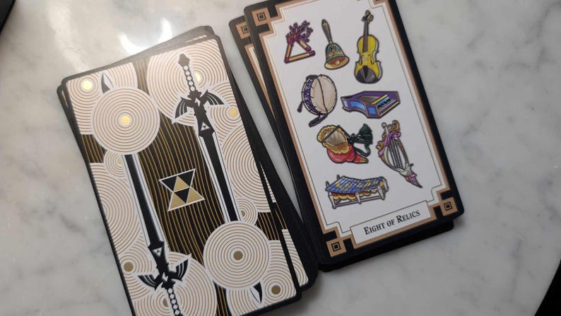 May include: Two tarot cards, one with a black and gold design featuring a sword and the other with a white background and colorful illustrations of musical instruments. The card with the musical instruments has the text "Eight of Relics".