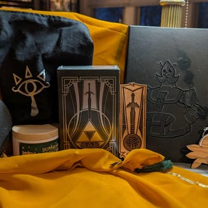 May include: A collection of themed merchandise, including a black drawstring bag with a white eye symbol, a slate coaster with a white "Z" and flower design, and a wooden cutout of a cartoon character. A yellow cloth is draped across the items.