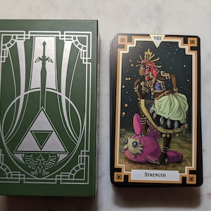May include: A green box with a silver design of a sword and a triangle. Next to the box is a tarot card with a black background and gold trim. The card depicts a woman in a colorful outfit standing on a pink creature. The card number VIII and the word "STRENGTH" are printed on the card.