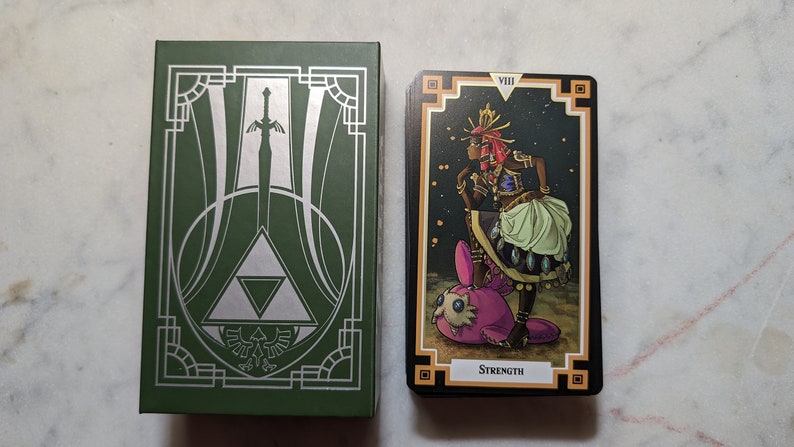 May include: A green box with a silver design of a sword and a triangle. Next to the box is a tarot card with a black background and gold trim. The card depicts a woman in a colorful outfit standing on a pink creature. The card number VIII and the word "STRENGTH" are printed on the card.