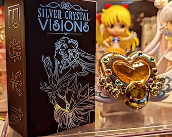 May include: A black rectangular box labeled Silver Crystal Visions with a silver line art illustration of a character. Beside it sits a gold heart-shaped accessory featuring a large amber-colored gem and smaller colorful embedded stones.