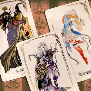 May include: Three tarot cards with gold trim and illustrations. The first card is the 9 of Swords, featuring a dark figure with horns and a cloak. The second card is the Page of Swords, featuring a figure in a blue and white outfit holding a sword. The third card is the 5 of Swords, featuring a figure with long white hair and a purple and black outfit.
