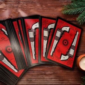 May include: A deck of playing cards with a red background and a white gun design. The cards are fanned out on a wooden surface, with a candle, pine cones, and greenery in the background. The cards have a vintage or antique aesthetic.