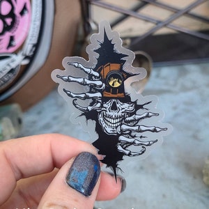 May include: A clear sticker with a black and white illustration of a skull wearing a miner's hat. The skull is peeking out from behind a pair of skeletal hands. The sticker has a distressed, ripped look.