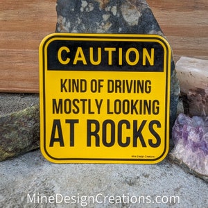 May include: A yellow and black square sign with rounded corners. The sign reads "CAUTION KIND OF DRIVING MOSTLY LOOKING AT ROCKS." The text "Mine Design Creations" is at the bottom. The sign is likely a humorous item for rock enthusiasts.