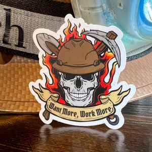 May include: A white sticker with a skull wearing a brown hard hat and holding two pickaxes. The skull is surrounded by flames and a banner that reads "Want More, Work More".