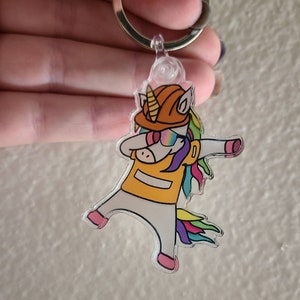 May include: A colorful acrylic keychain featuring a unicorn wearing a construction worker hat, sunglasses, and a yellow vest. The unicorn is in a dabbing pose.