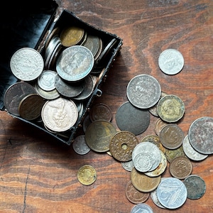 May include: A black treasure chest overflowing with a collection of silver and gold coins. The chest is open and the coins are spilling out onto a wooden surface.