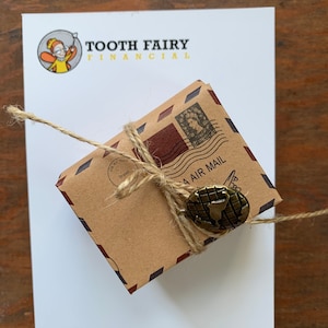 May include: A small brown package wrapped in brown paper with a twine bow and a globe-shaped metal charm. The package has a red and white airmail stamp and the text "AIR MAIL" printed on it. The package is sitting on a white notepad with the text "TOOTH FAIRY FINANCIAL" printed on it.