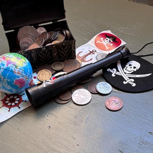 May include: A black plastic telescope with a pirate sticker, a small open treasure chest filled with metallic coins, a miniature globe, several circular pirate-themed stickers featuring skulls and anchors, and a black fabric patch with a white skull and crossbones.