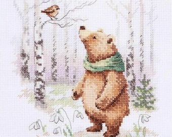 May include: A cross-stitch embroidery pattern on white fabric featuring a brown bear wearing a green scarf standing in a forest of birch trees. A small brown bird sits on a branch above the bear, and white snowdrop flowers grow at the base.