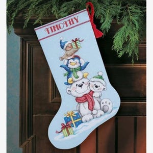 Peut inclure: Une chaussette de Noël bleu clair avec le nom TIMOTHY imprimé en haut. Le motif représente une pile d'animaux incluant un oiseau, un manchot et un ours polaire avec un ourson, tous portant des accessoires d'hiver et tenant des cadeaux.