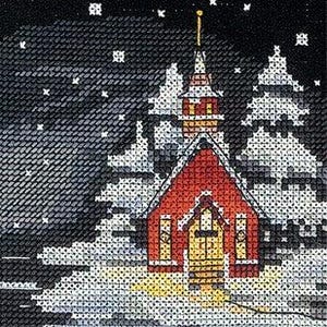 Op de afbeelding: Een kruissteekborduurwerk met een rode kerk met gele ramen, omringd door besneeuwde dennenbomen tegen een zwarte nachtelijke hemel vol witte sterren. De scène bevat een reflectie van de kerk op een donker, ijzig oppervlak onderaan.