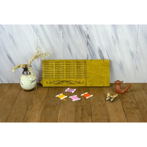 Puede incluir: Un organizador de hilos rectangular de madera color amarillo mostaza con ranuras horizontales y un panel sólido, acompañado de un jarrón de cerámica con trigo seco, un pequeño silbato de barro con forma de pájaro, un adorno de mariposa y cuatro bobinas.