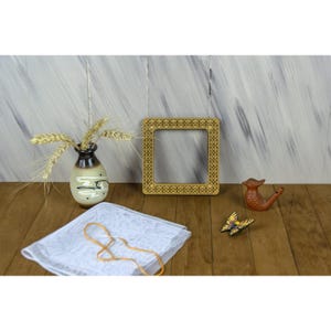 Può includere: Una cornice quadrata in legno con un motivo geometrico intagliato, un piccolo vaso in ceramica contenente spighe di grano, un fischietto in terracotta a forma di uccello, un ornamento a farfalla giallo e nero e un quadrato di tessuto bianco.