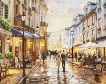 May include: A cross-stitch pattern depicting a European street scene with multi-story buildings, outdoor cafe seating, street lamps, and pedestrians walking along a cobblestone path. The scene features warm lighting, flower boxes, and umbrellas.