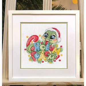 Puede incluir: Un bordado en punto de cruz enmarcado que presenta una serpiente de dibujos animados verde con un gorro de Papá Noel rojo y blanco. La serpiente está rodeada de agujas de pino verdes, bayas rojas y puntos amarillos. El texto cirílico "С НОВЫМ ГОДОМ!" está bordado en letras coloridas.