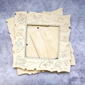 May include: A light-colored wooden square frame with irregular, map-like edges. The frame features laser-engraved illustrations including a compass, a castle, a sailing ship, palm trees, a magnifying glass, a volcano, and sea creatures.