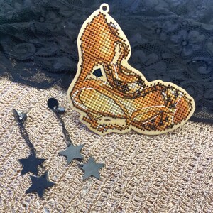 May include: A wooden ornament featuring a cross-stitched high-heeled shoe in shades of orange, yellow, and brown, paired with a set of dangling earrings consisting of black metal chains and two matte black star-shaped charms on each earring.