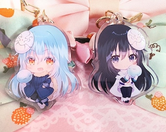 以下が含まれることがあります： 白い背景に、青いキャラクターと黒いキャラクターの2つのアニメキャラクターチャーム。両方のキャラクターが小さな青い球を持っています。チャームは金色のチェーンに取り付けられています。