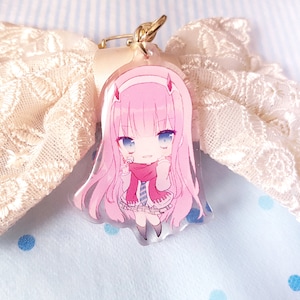May include: A pink and white anime character keychain with a red scarf and blue and white striped shirt. The character has pink hair and blue eyes.