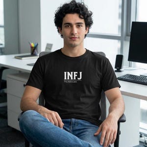 May include: Black t-shirt with the white text "INFJ The Advocate". The shirt is a crew neck style. The person in the image is wearing blue jeans and is seated in a chair.