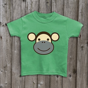 May include: A green t-shirt with a cartoon monkey face on the front. The monkey has brown ears, a yellow face, and a gray mouth.