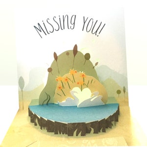 May include: A pop-up card with a 3D scene of two white swans swimming in a blue lake surrounded by green reeds and yellow flowers. The card has the words "Missing You!" written on the front.