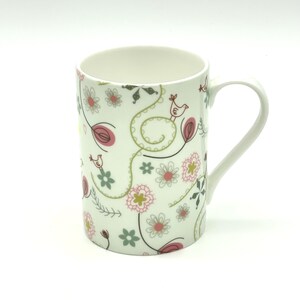 May include: White ceramic mug with a floral pattern in shades of pink, green, and yellow. The pattern features stylised flowers, leaves, and birds.