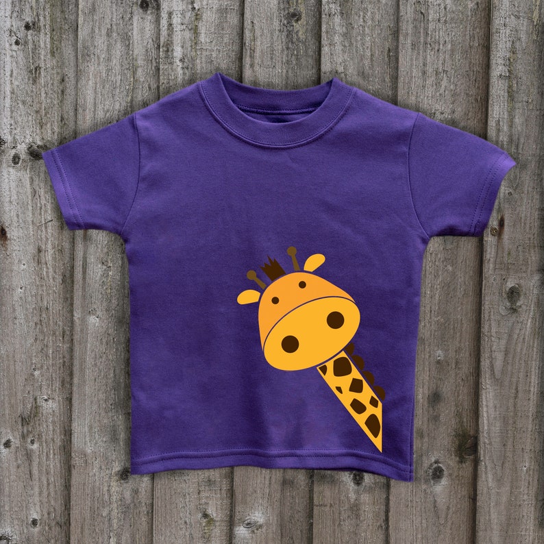 May include: Purple short-sleeved t-shirt with a cartoon giraffe peeking out from the bottom right corner. The giraffe is orange and brown with a light brown outline.