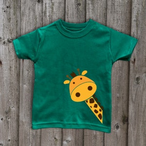 May include: A green t-shirt with a cartoon giraffe peeking out from the bottom left corner. The giraffe is yellow with brown spots and has a friendly expression.