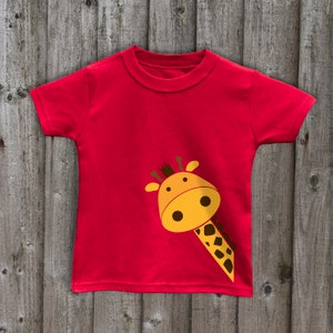 May include: Red short-sleeved t-shirt with a yellow giraffe head peeking out from the side.