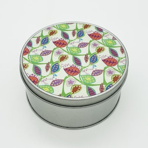 May include: A circular, silver-coloured tin with a lid displaying a vibrant floral pattern. The lid's design incorporates green vines, diverse leaf shapes in blue, orange, and purple, and small purple flowers set against a white backdrop. The tin is likely for storage.