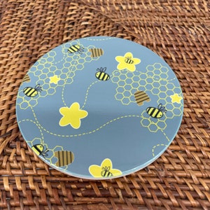 Puede incluir: Un posavasos redondo gris con un patrón de panal y flores y abejas amarillas. El posavasos tiene un fondo azul claro con un contorno blanco alrededor del diseño.