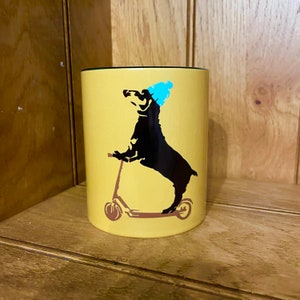 Puede incluir: Una taza de cerámica amarilla con un borde negro presenta una silueta negra de un dachshund con un gorro azul y conduciendo un scooter.