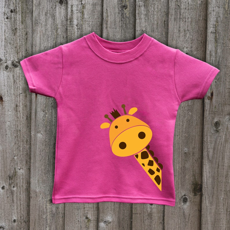 May include: A pink t-shirt with a cartoon giraffe peeking out from the side. The giraffe is yellow with brown spots and has a brown nose and eyes.