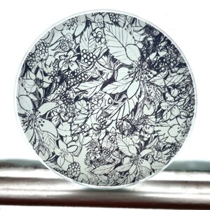 May include: A round, white plate with a black and white floral design. The design features leaves, flowers, and berries.