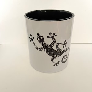 Puede incluir: Taza de cerámica blanca con borde negro y diseño de gecko estampado en blanco y negro.