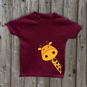 May include: A maroon t-shirt with a cartoon giraffe peeking out from the bottom right corner. The giraffe is yellow with brown spots and has a friendly expression.