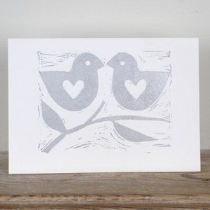 A hand printed greeting card by artist Melissa Birch featuring two birds with love heart symbols in silver ink on a white background