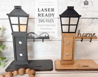 May include: Two laser-cut wooden lamppost designs with hooks for hanging keys or other items. One lamppost is black with a white light, the other is brown with a white light. Both lampposts have the word "home" above the hooks. The text "LASER READY SVG FILES" is in the upper left corner of the image. The text "Instant Download Instructions Commercial Use" is below the text "LASER READY SVG FILES". The text "2 DESIGN OPTIONS!" is at the bottom of the image.