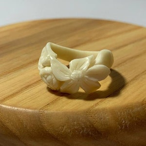 Puede incluir: Un anillo blanco tallado con un diseño floral. El anillo está hecho de hueso y presenta tres flores.