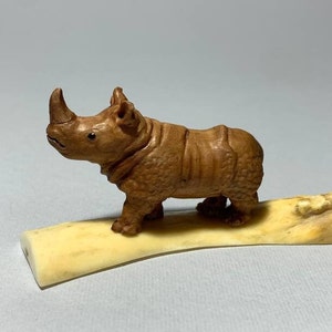 May include: A detailed wooden carving of a rhinoceros. The rhino is brown with textured skin and a prominent horn. It stands on a cream-colored, curved base, showcasing the craftsmanship of the sculpture.